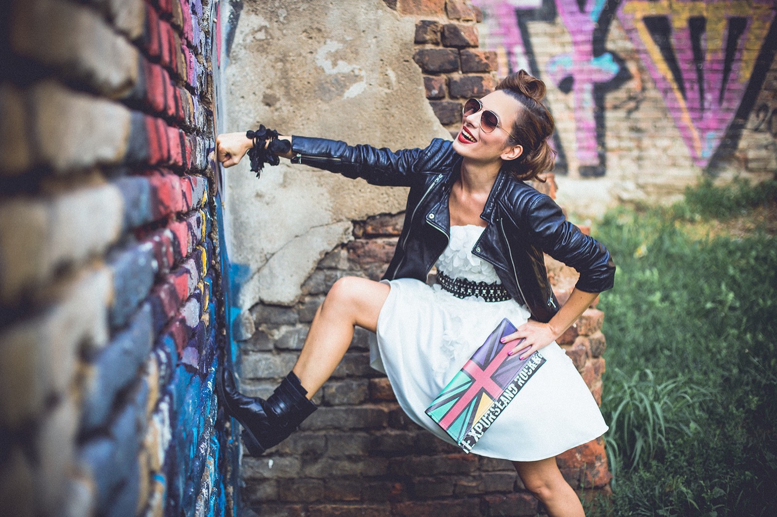 Ona se smeje uz grafitni zid — kožna jakna, bela haljina i Union Jack torba
