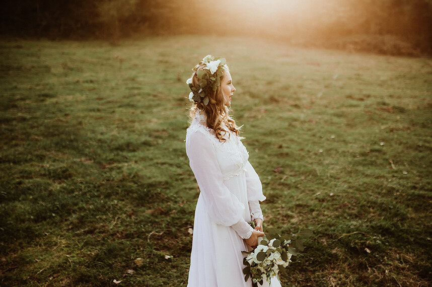 Mlada Paula stoji na livadi pri zalasku sunca u maminoj beloj haljini sa venčićem od cveća na glavi, drži buket iza leđa