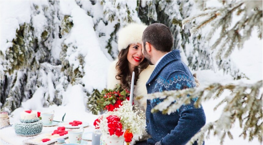 Par za stolom u snegu — mlada sa crvenim usnama, bela krznena kapa, mladoženja u plavom nordijskom džemperu